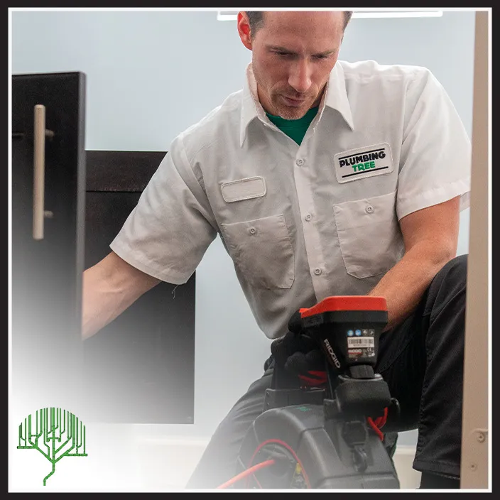 A plumber wearing a "Plumbing Tree" shirt uses a diagnostic tool to inspect plumbing inside a building.