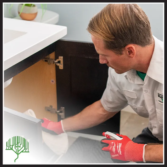 A person wearing gloves and a uniform cleans the inside of a bathroom cabinet with a cloth and spray bottle. A sink and plant are visible in the background.