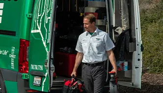 A technician in uniform carries tools while walking away from an open green service van filled with equipment.