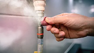 A hand touching a leaking pipe with steam or vapor escaping from a fitting in an indoor setting.