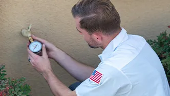 A man in a white shirt with a US flag patch checks a water pressure gauge attached to an outdoor faucet.