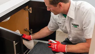 A man wearing red gloves and a uniform cleans under a kitchen sink cabinet with a cloth and spray bottle.