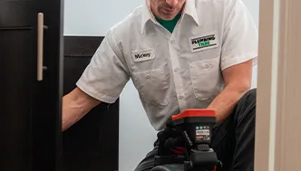 A technician in a uniform labeled "Vicky" uses inspection equipment while kneeling next to a piece of furniture indoors.