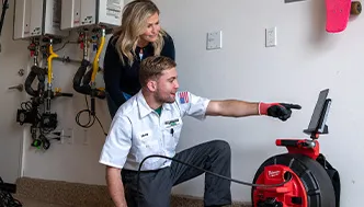 A technician kneels while explaining information on a screen to a woman in a utility room with equipment on the wall.