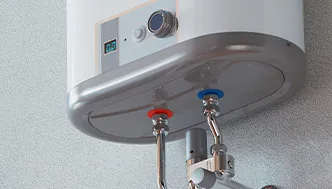 Close-up view of an electric water heater mounted on a wall, showing two pipes with red and blue valve indicators for hot and cold water connections.