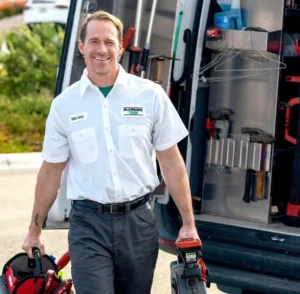 A plumber in a white uniform shirt labeled "Mickey" stands beside a work van, holding plumbing tools and equipment, with the van door open showing organized tools inside.