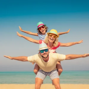 Three people wearing hats and sunglasses pose on a beach, stacked on each other’s backs with arms outstretched, in front of the sea and blue sky.