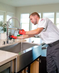 A plumber wearing red gloves and a white shirt inspects a kitchen faucet while water flows into a stainless steel sink.